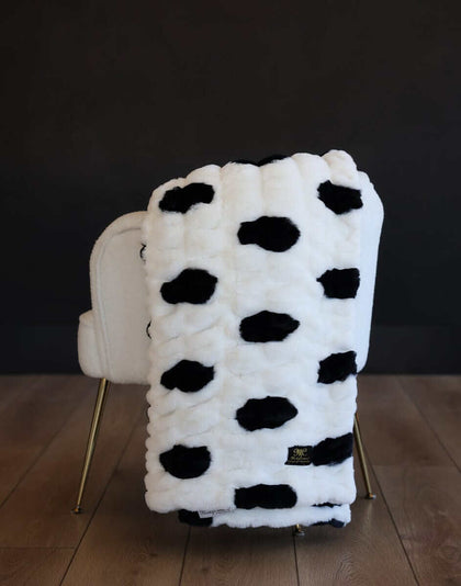 White Minky Hugs Polka Dot Marshmallow plush blanket with black spots draped over a cream chair.