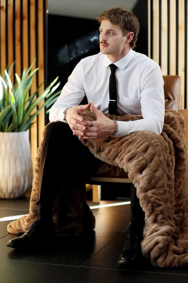 Man sitting in a chair wrapped in the Biggest Hugs Milk Chocolate weighted blanket, showcasing its cozy warmth and soft texture.