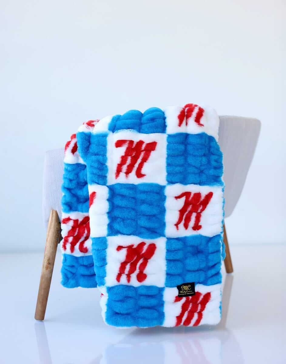 Ole Miss checkerboard throw blanket in blue and white with red “M” logos, draped over a light chair.
