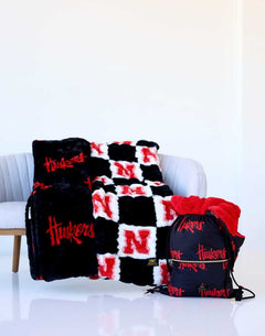 Nebraska Huskers throw blankets in black, red, and white displayed on a chair with a matching drawstring bag.