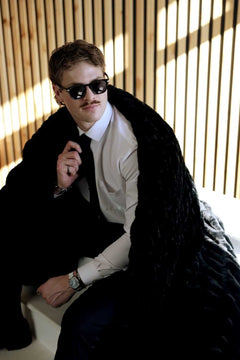 Model wearing Walker Kessler Biggest Hugs Black Blanket, sitting stylishly with sunglasses, in a modern setting.