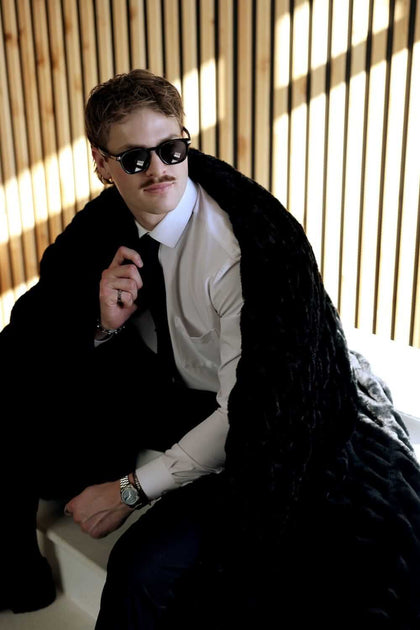 Model wearing Walker Kessler Biggest Hugs Black Blanket, sitting stylishly with sunglasses, in a modern setting.