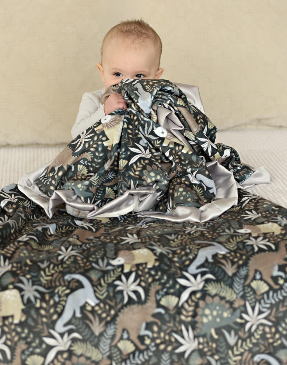 Little Dino Gray Blanket on a baby, featuring playful dinosaurs and a soft satin ruffle.