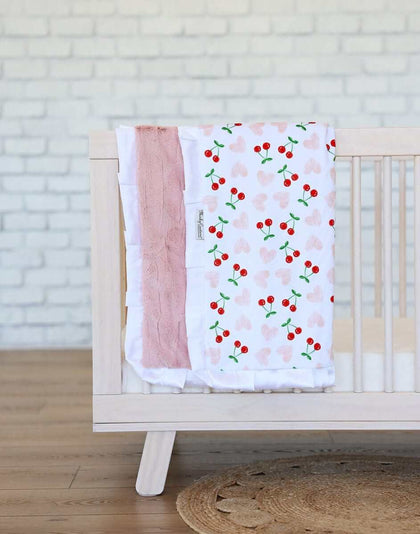 Minky Couture Cheery Cherry Blanket draped over a crib featuring cherry designs and a plush apricot backing.