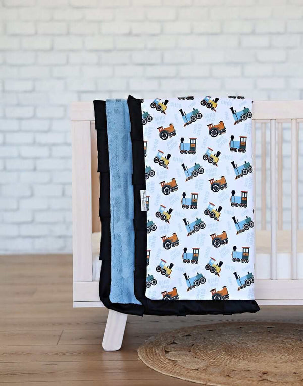 Minky Couture Choo Choo Blanket hanging on a crib, featuring colorful train patterns and a black satin ruffle.