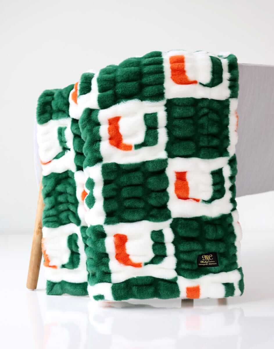 Green, white, and orange Miami Hurricanes Minky Couture blanket folded and draped over a chair, showing its textured pattern.