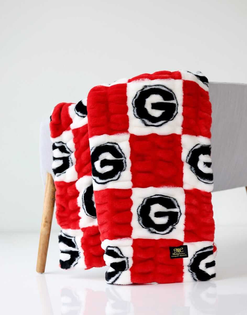 Red and white Georgia Hugs Minky Couture blanket folded and draped over a chair, showing its textured logo pattern.