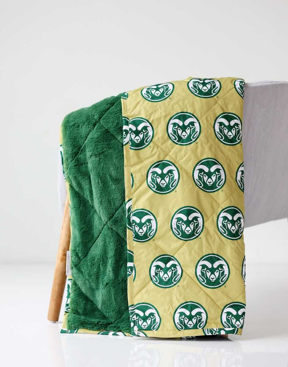 Colorado Rams Everywhere Minky Couture blanket folded over a chair, showing gold logo side and green plush lining.