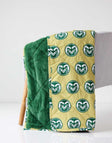 Colorado Rams Everywhere Minky Couture blanket folded over a chair, showing gold logo side and green plush lining.