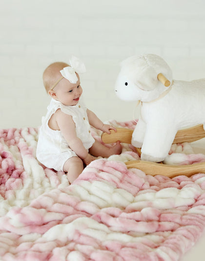 Baby sitting on a pink cow print Hugs Dollie Blanket, playing with a plush toy sheep.