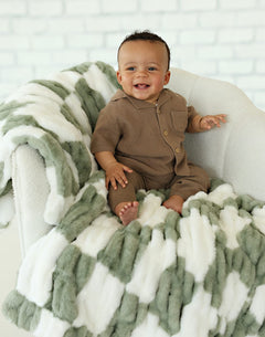 Happy baby sitting on a Hugs Check Sage Blanket in a cozy setting.
