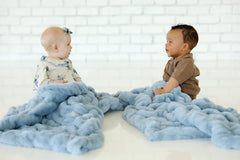 Two babies sitting on a floor each wrapped in the Hugs Alaskan Blue Blanket, showcasing its plush texture and calming color.