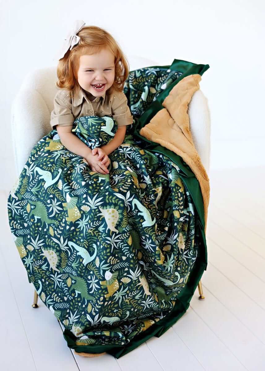 Minky Couture Little Dino Green Blanket featuring colorful dinosaurs with a child happily wrapped in it.