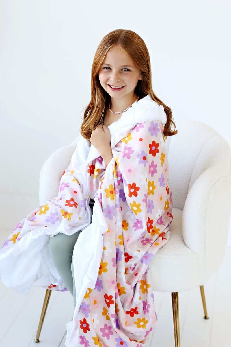 A girl wrapped in a vibrant Flower Power Blanket with floral design and white plush backing, smiling in a cozy setting.