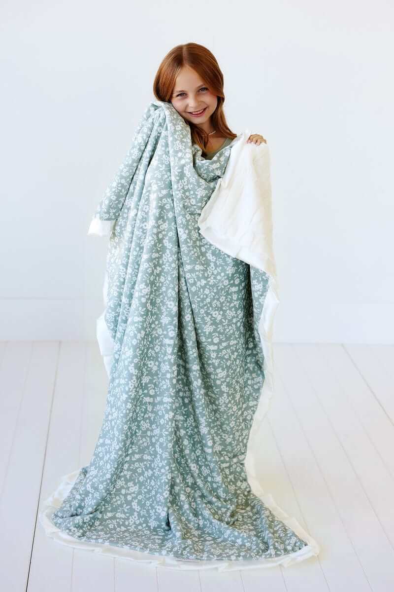 A girl smiling while wrapped in the Dainty Floral Sage Blanket, showcasing its floral pattern and cream satin ruffle.