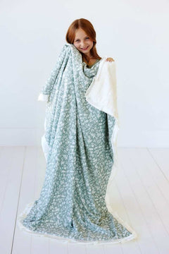 A girl smiling while wrapped in the Dainty Floral Sage Blanket, showcasing its floral pattern and cream satin ruffle.