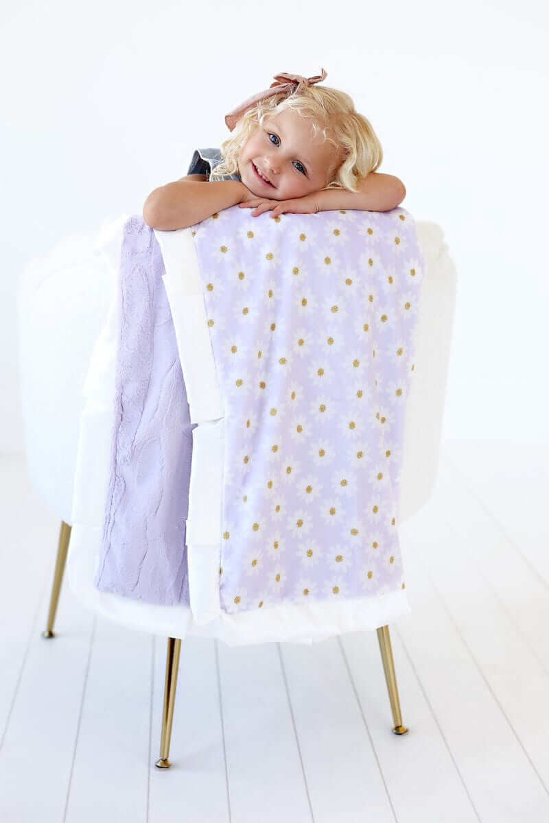 Cheerful child resting with the Lavender Daisy Blanket draped on a chair, showcasing its daisies and lilac color.