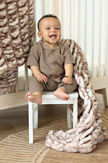 Happy baby wearing brown outfit sitting on a chair beside the cozy Hugs Topaz Blanket.
