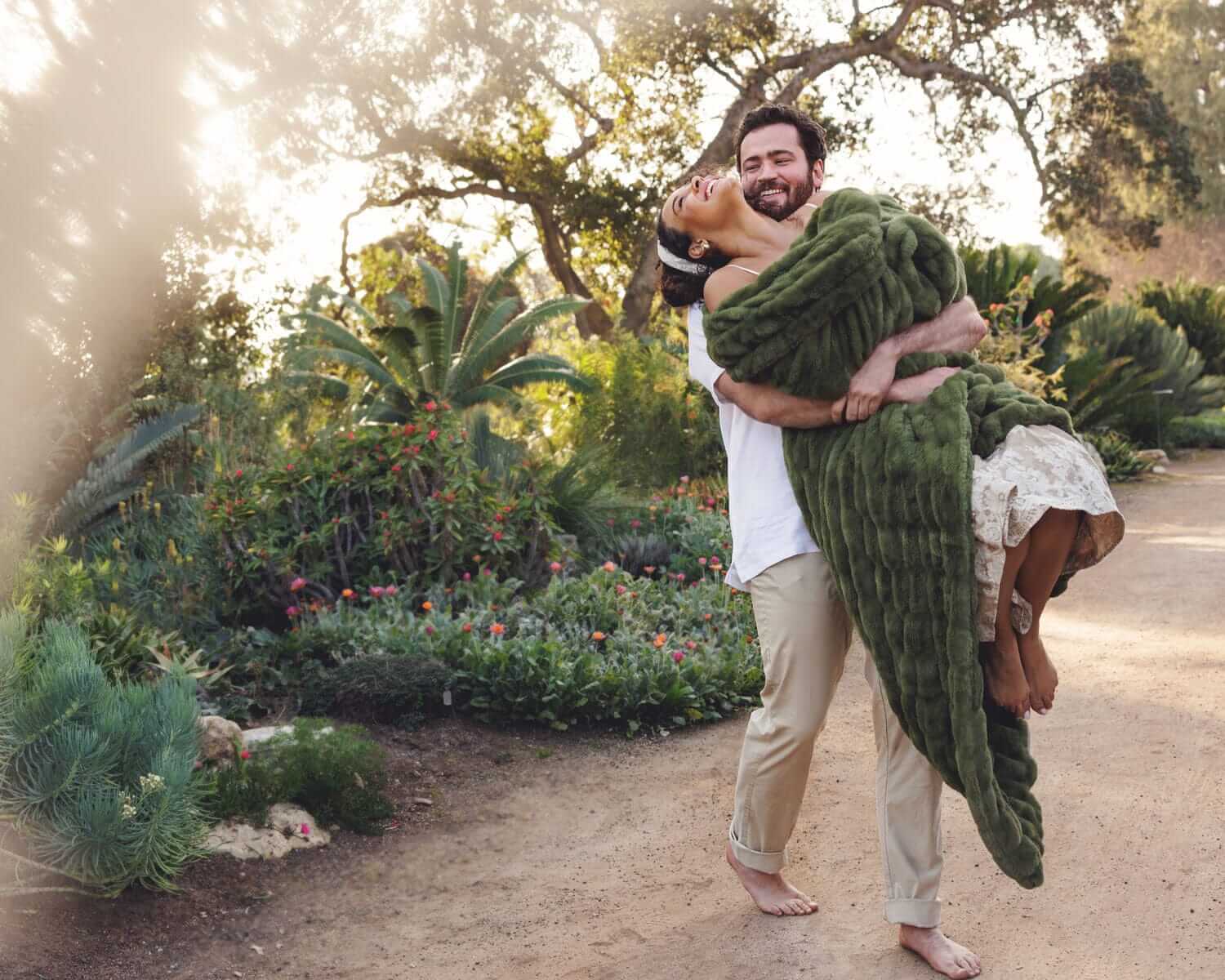 Man holding a woman wrapped in the Hugs Camp Green Blanket in a beautiful outdoor garden setting.
