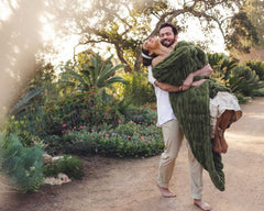 Man holding a woman wrapped in the Hugs Camp Green Blanket in a beautiful outdoor garden setting.