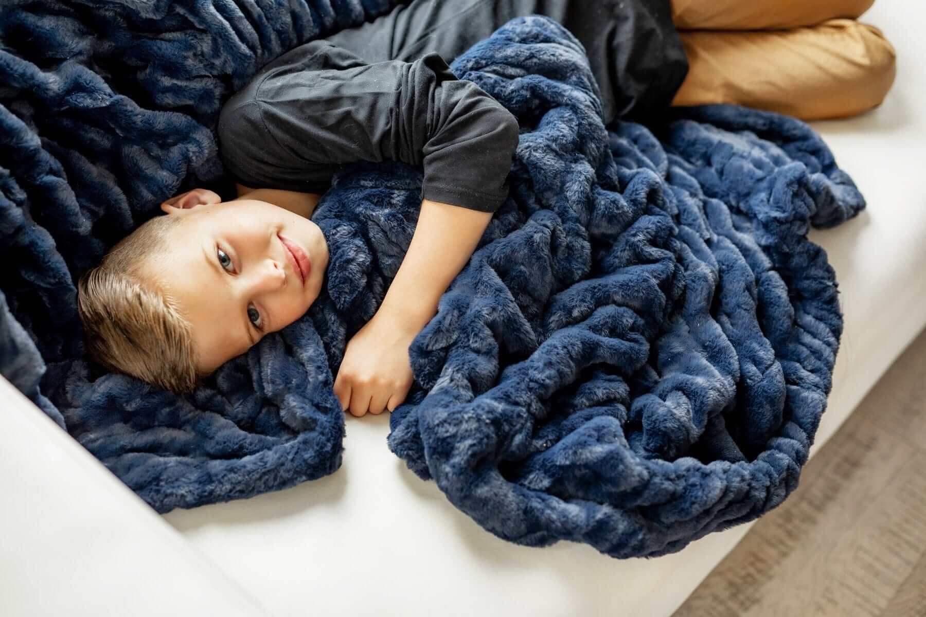 Child smiling while cuddled in a navy blue Minky Couture weighted blanket with a thick, plush braided texture.