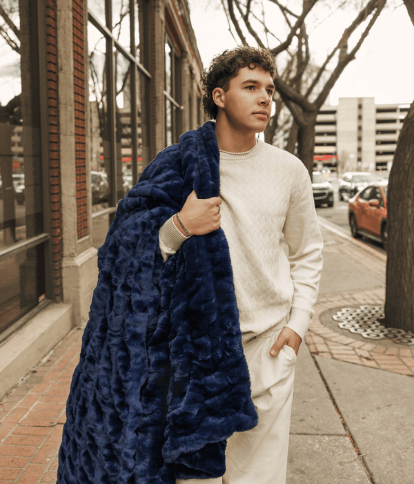 A model wraps the Biggest Hugs Navy Blanket around himself while walking down the street, showcasing its comfort and style.