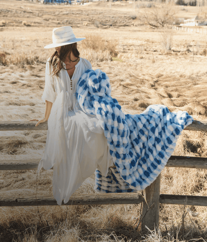 Woman in a white dress with blue and white throw blanket sitting on a fence in a serene outdoor landscape.