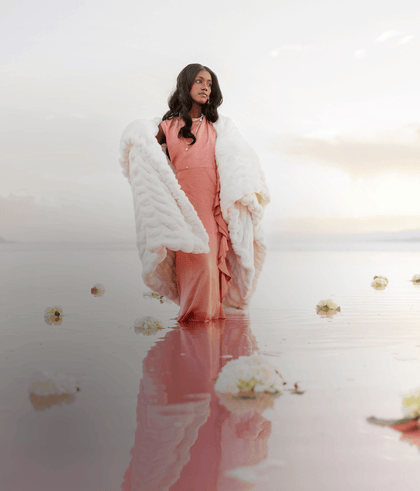 Woman in a cozy white blanket standing on a serene, reflective lake surrounded by flowers and a colorful sky.