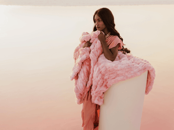 A woman draped in a fluffy pink blanket sits peacefully by the serene waters at sunset.