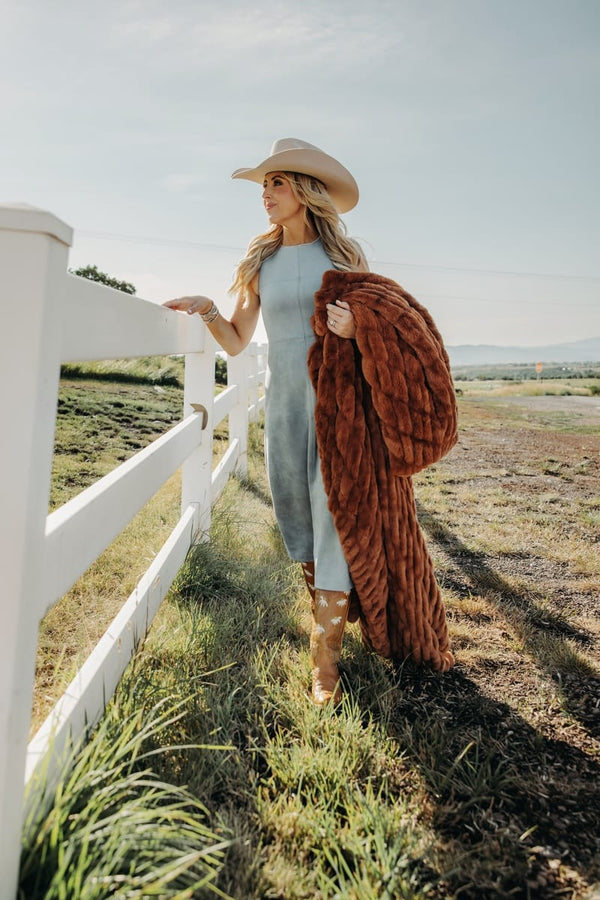 A woman in a light blue dress and cowboy hat stands outdoors beside a white fence, holding a rust-brown throw blanket in a sunny field.