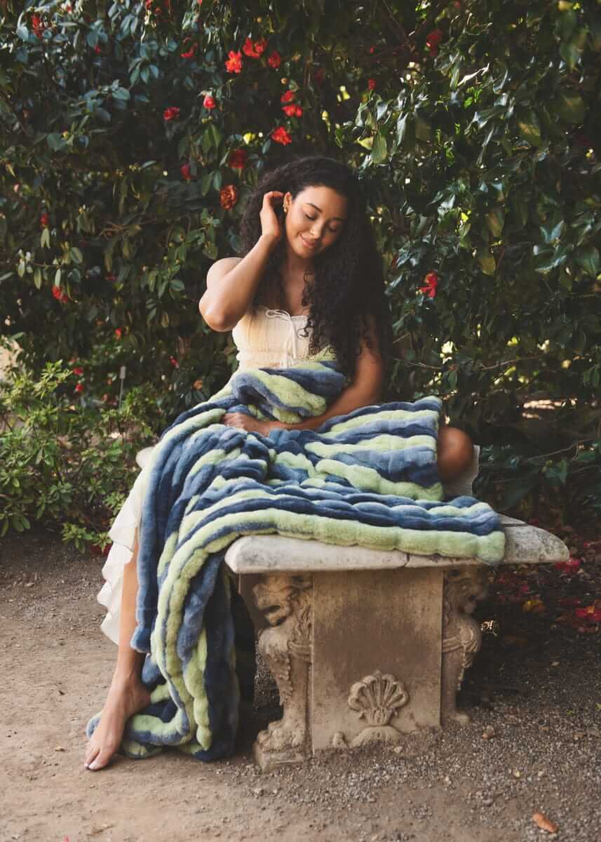 Hugs Bold Stripe Blanket draped over a woman sitting on a stone bench surrounded by vibrant flowers.