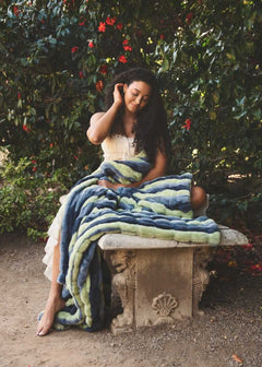 Hugs Bold Stripe Blanket draped over a woman sitting on a stone bench surrounded by vibrant flowers.