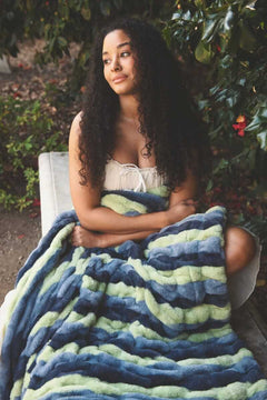 Young woman wrapped in Minky Couture Hugs Bold Stripe Blanket sitting outdoors amongst greenery.