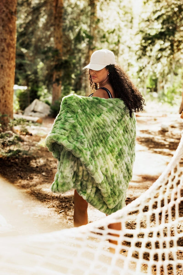 A woman standing in a sunlit forest clearing, wrapped in a chunky green faux-fur style throw blanket and wearing a white baseball cap, with a white hammock in the foreground.