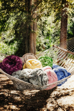 A collection of plush, chunky knit blankets in various colors, including purple, yellow, green, pink, blue, and gray, arranged in a woven hammock suspended outdoors between trees.