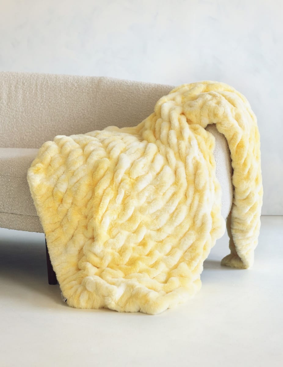 A chunky, stretchy-textured throw blanket in pale yellow and cream draped over a light beige sofa in a minimalist indoor setting.