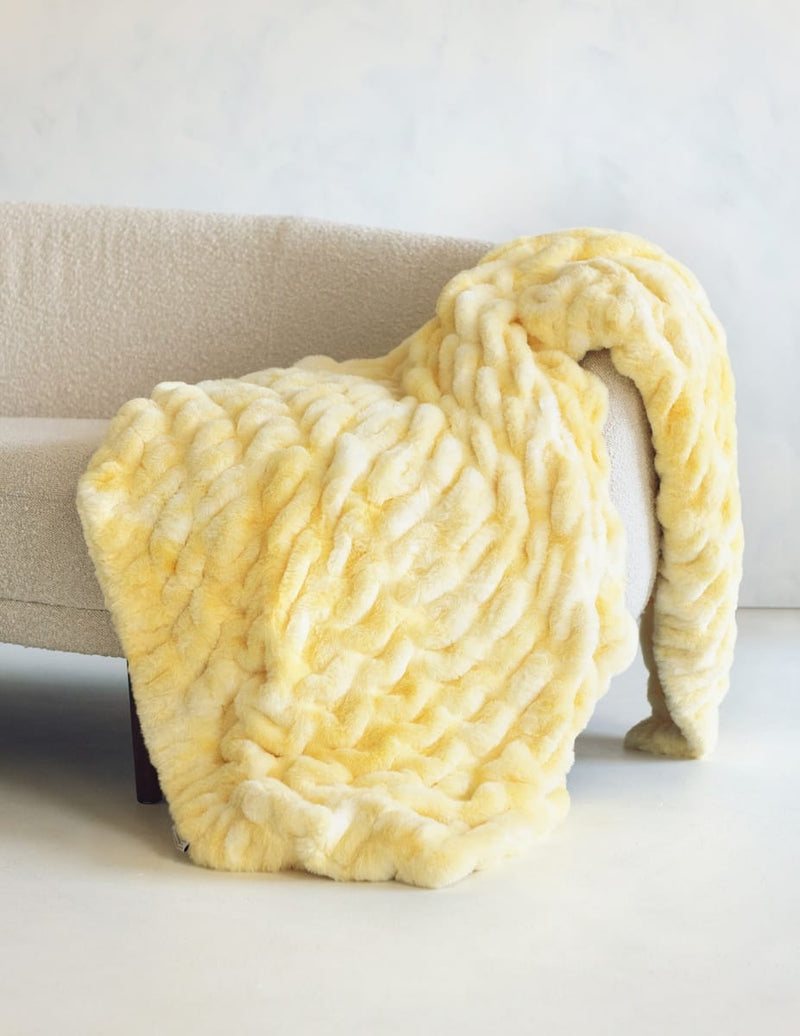 A chunky, stretchy-textured throw blanket in pale yellow and cream draped over a light beige sofa in a minimalist indoor setting.