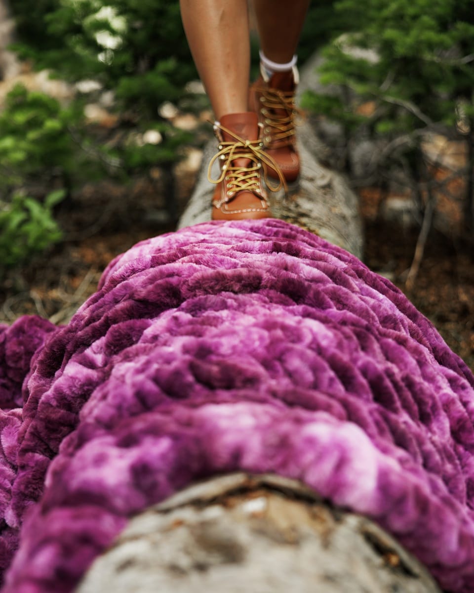 A close-up of a plush, textured purple blanket draped over a log in a forest, with a person's legs and brown lace-up boots visible stepping across the log above.