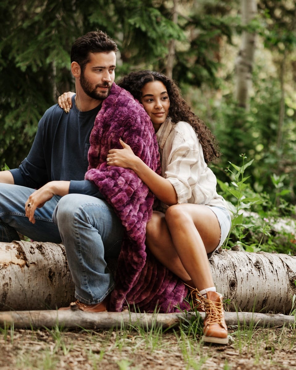 A man and a woman sit together on a birch log in a forest, wrapped in a plush, textured magenta-purple throw blanket. 