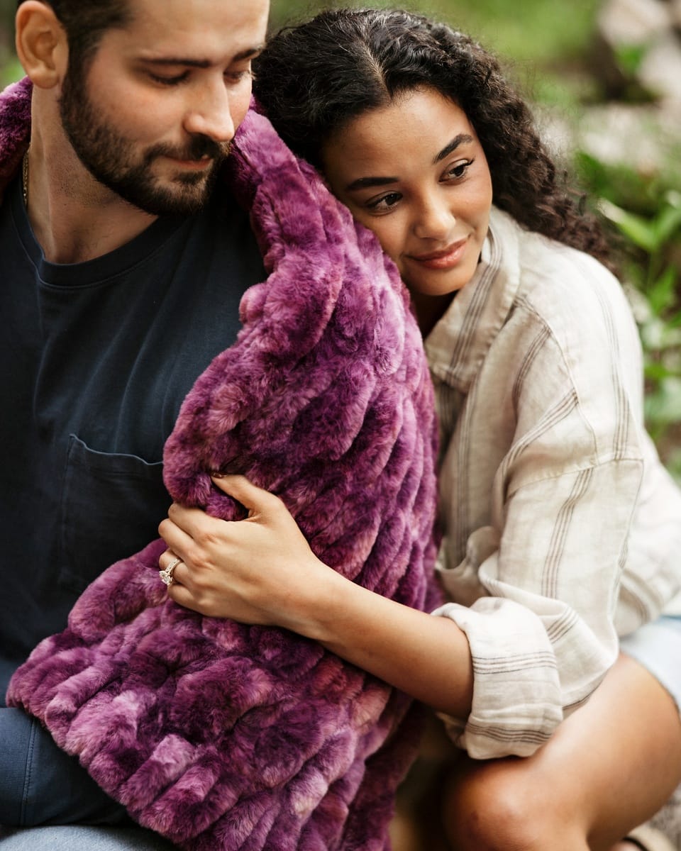 A close-up lifestyle shot of a couple hugging, with the woman wrapped in a plush, textured purple throw blanket outdoors.