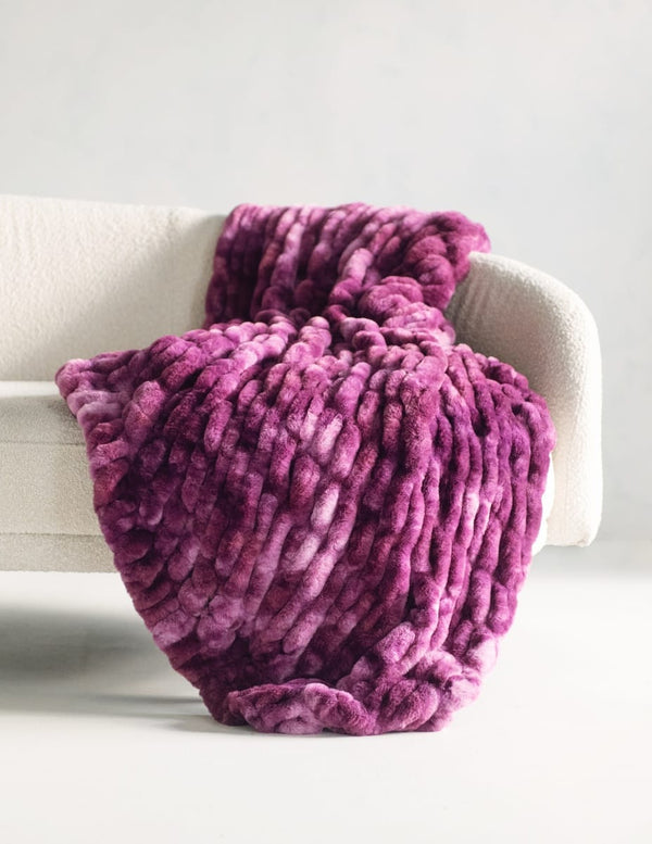 A chunky, stretchy throw blanket in a rich magenta-purple color, draped over a modern white armchair against a clean, light gray background.