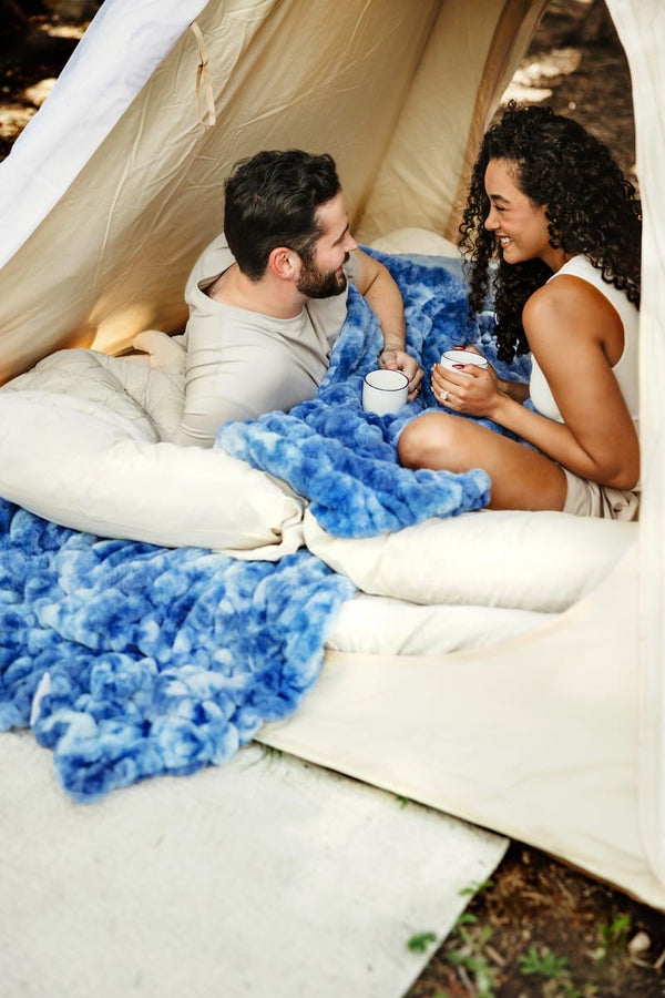A couple relaxes inside a beige canvas glamping tent, sharing a blue fuzzy blanket and mugs, with a soft white pillow in the foreground.