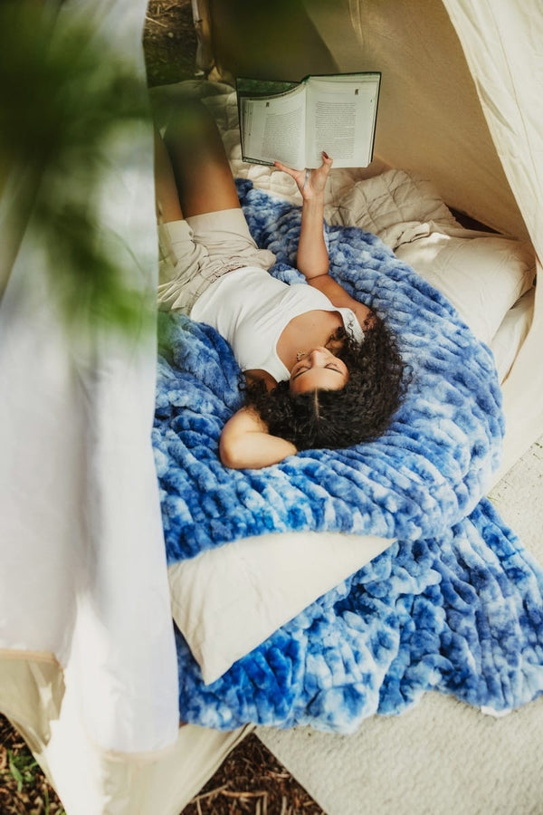 A woman lies on her back inside a beige tent, reading a book while wrapped in a chunky, textured blue throw blanket with white pillows.