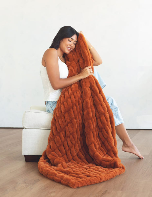 A woman sitting on a white armchair holds up a stretchy blanket in a warm rust-orange color against a plain white wall and light wood floor.