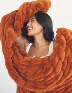 A woman wrapped in a chunky, oversized rust-orange faux fur blanket, smiling against a plain light background.