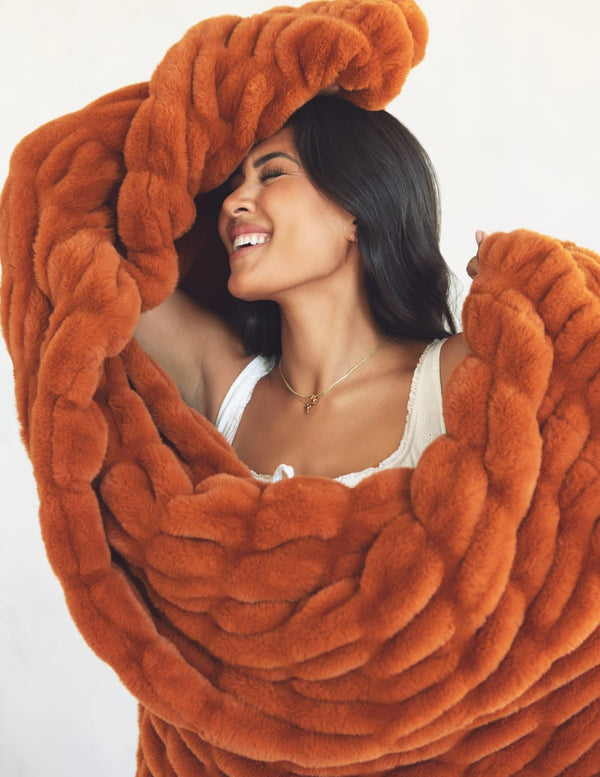 A woman wrapped in a chunky, oversized rust-orange faux fur blanket, smiling against a plain light background.
