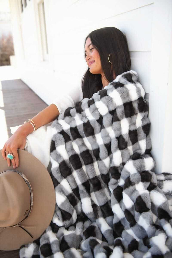 Young woman sitting leaning against a white house with a Hugs Gingham Charcoal Blanket.