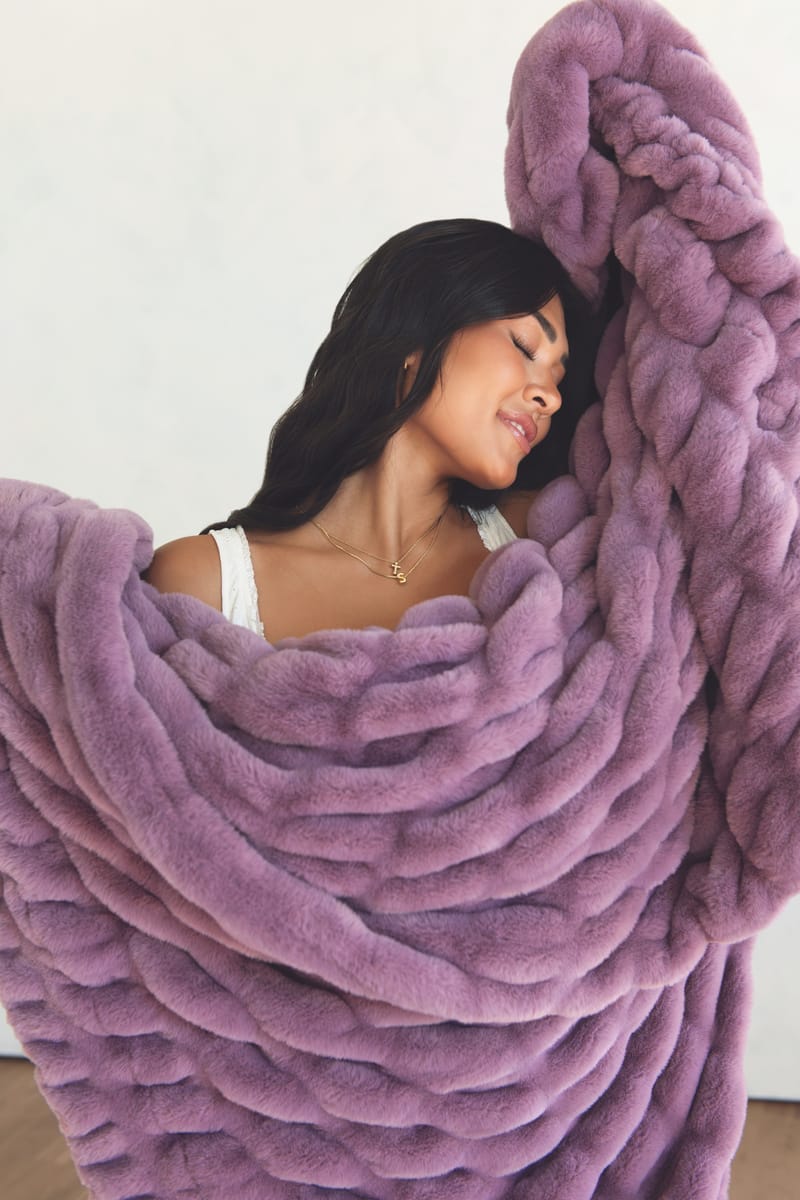 A woman wrapped in a plush throw blanket in a dusty lavender-purple color, set against a plain off-white wall.
