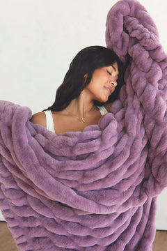 A woman wrapped in a plush throw blanket in a dusty lavender-purple color, set against a plain off-white wall.