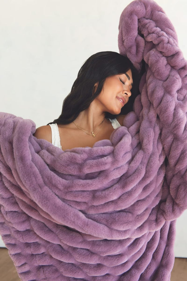 A woman wrapped in a plush throw blanket in a dusty lavender-purple color, set against a plain off-white wall.